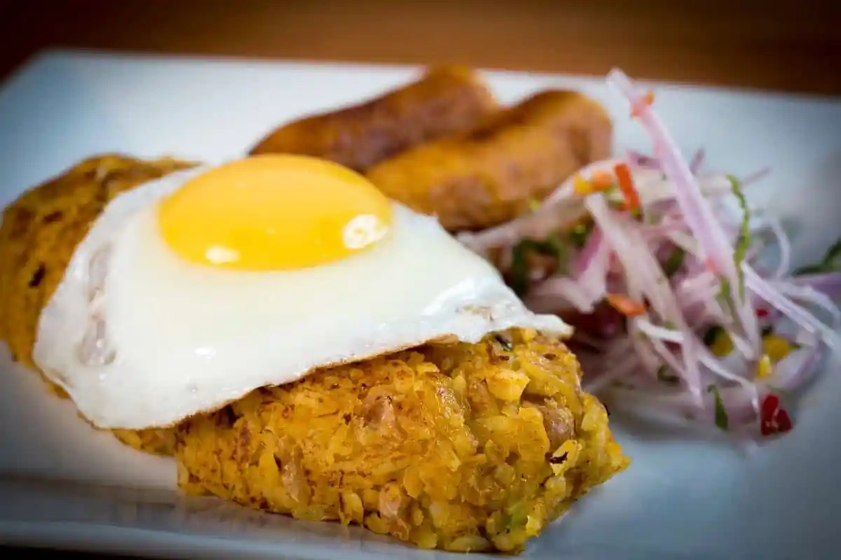 Tacu tacu con huevo frito encima, acompañado de plátano dorado y salsa criolla con cebolla roja