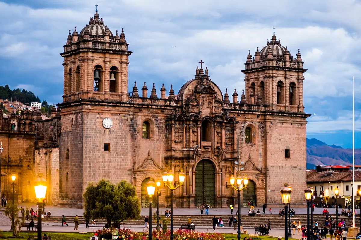 Fachada de la Catedral del Cusco al atardecer, con sus dos campanarios, portada barroca y faroles encendidos en la Plaza de Armas
