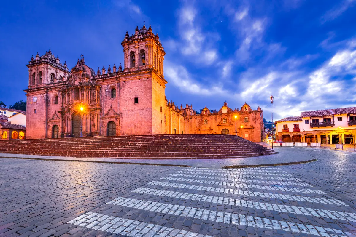 Catedral del Cusco iluminada durante la hora azul, con la Iglesia del Triunfo a un lado, escalinatas de piedra y cielo con nubes
