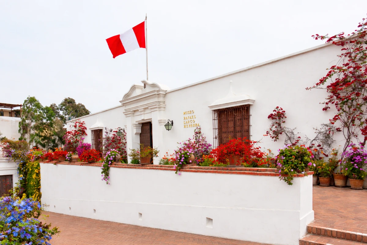 Vista exterior del Museo Larco en Pueblo Libre, con su casona virreinal blanca, flores en los jardines y la bandera del Perú sobre la entrada.