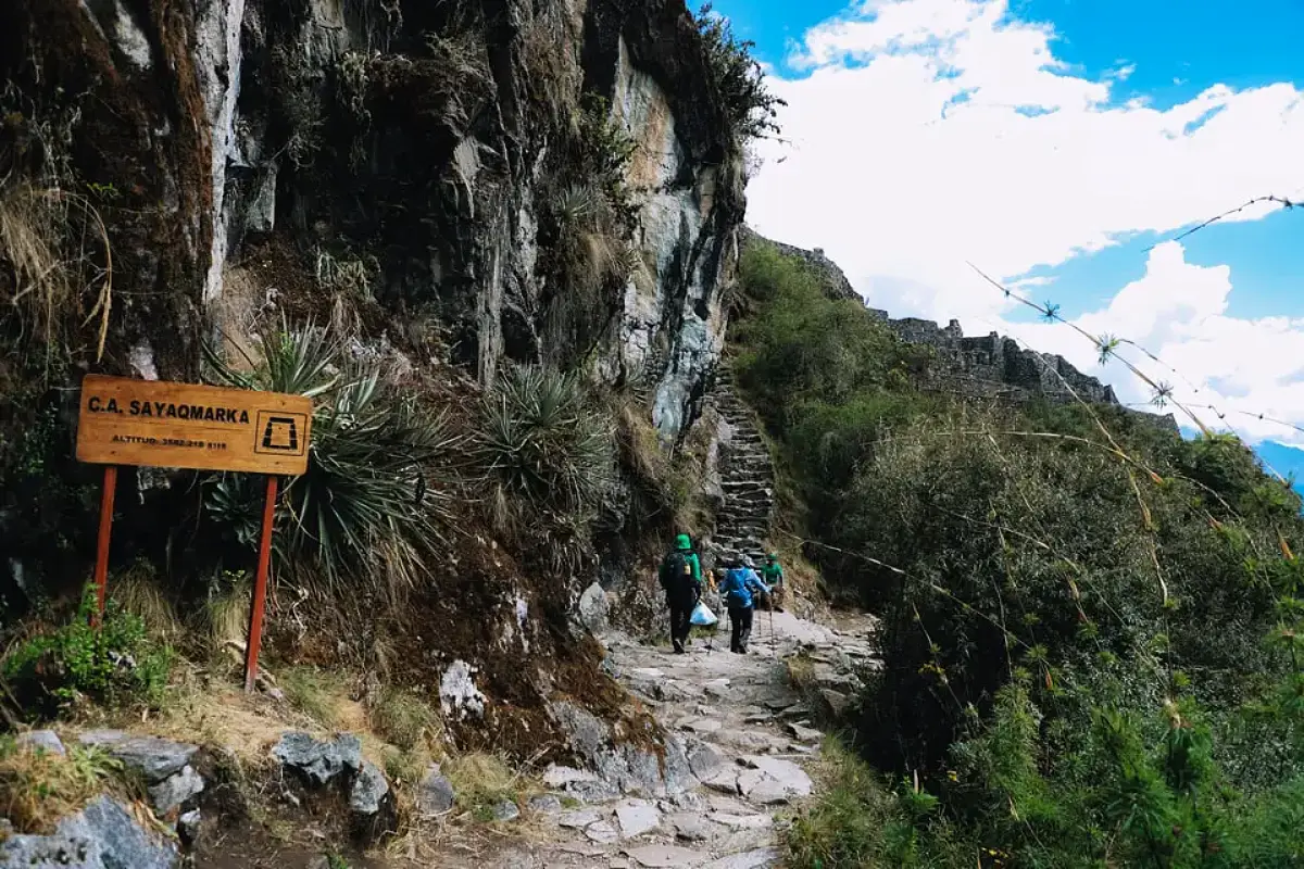 Viajeros junto al letrero de Sayacmarca, frente a una escalinata de piedra del Camino Inca